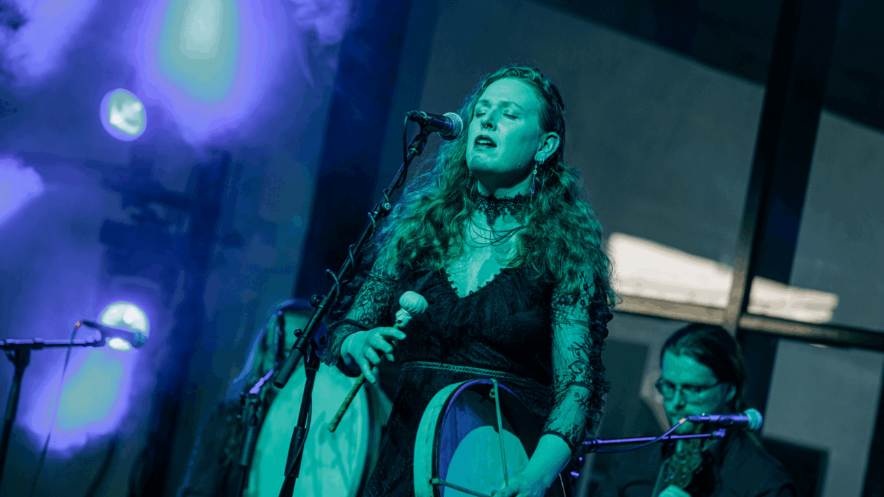 Women singing into a microphone on stage, with 2 musicians sitting behind her.