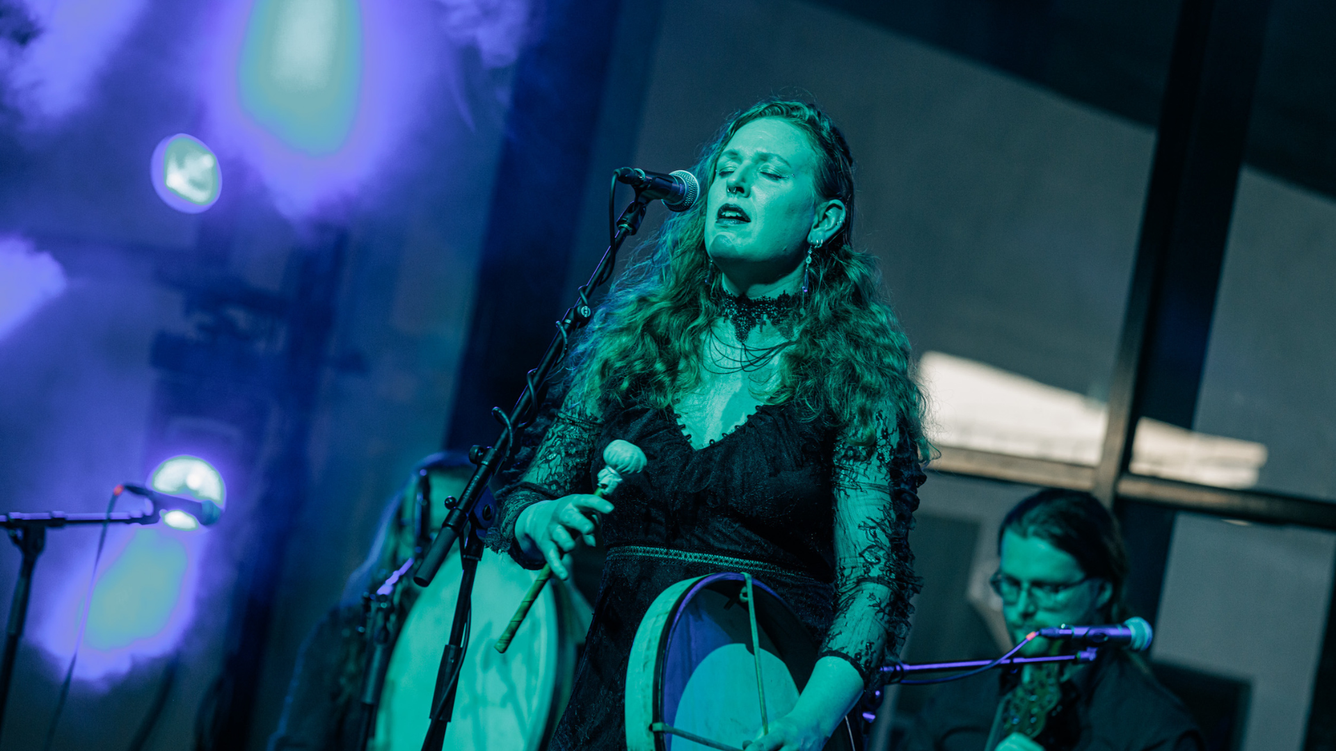 Women singing into a microphone on stage, with 2 musicians sitting behind her.