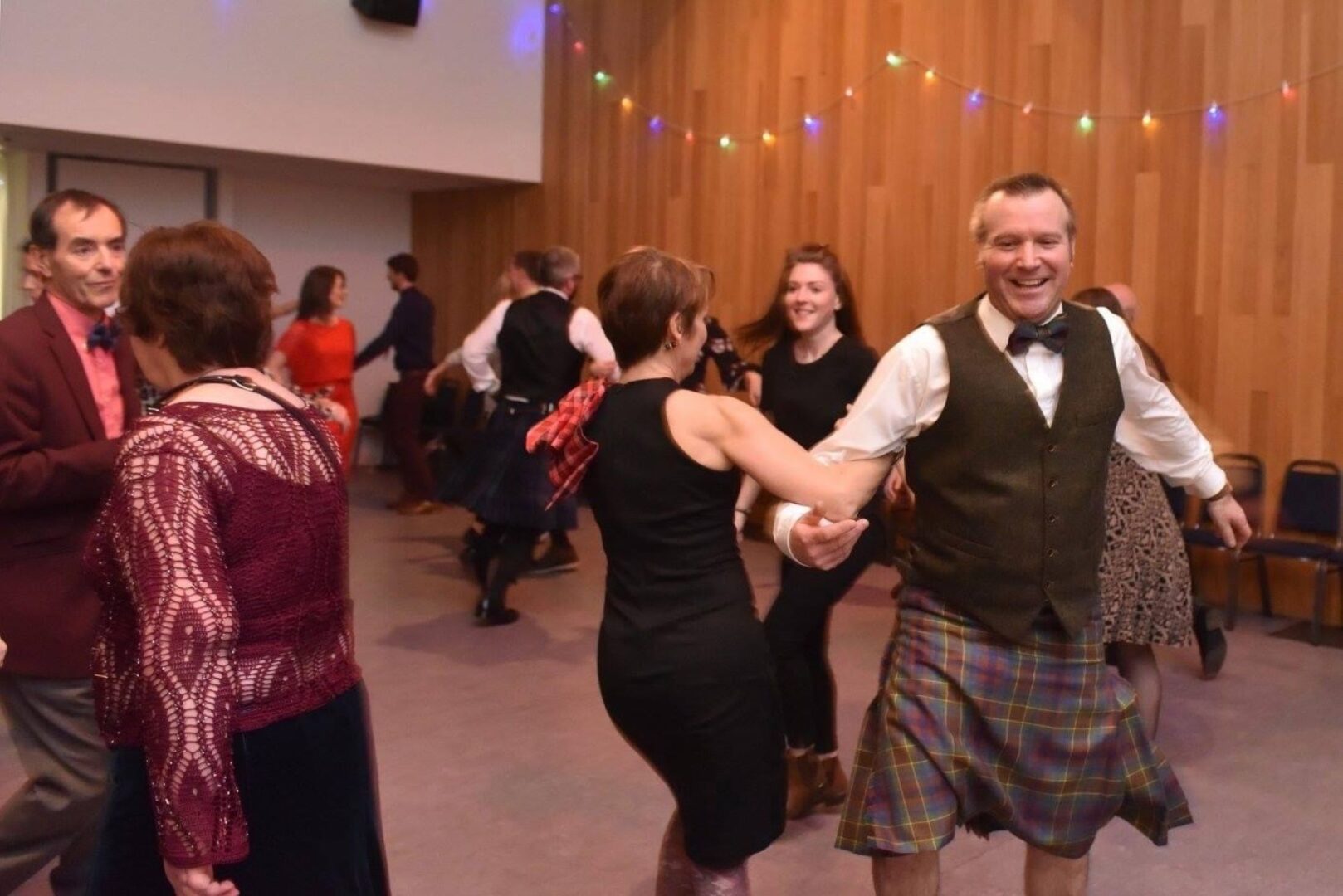 Group of people dancing in pairs. Some of the men are wearing traditional Scottish kilts.