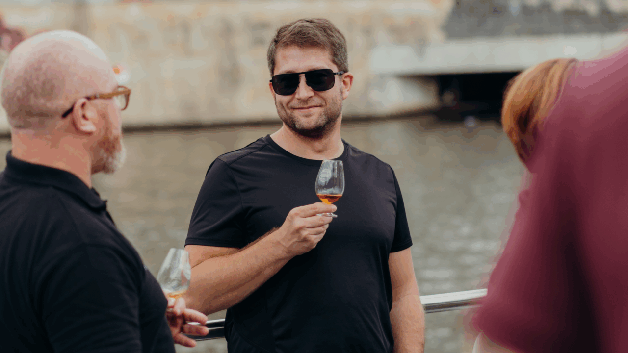 Man with dark sunglasses holding a filled whisky glass, standing on a boat. There are 2 people beside him.