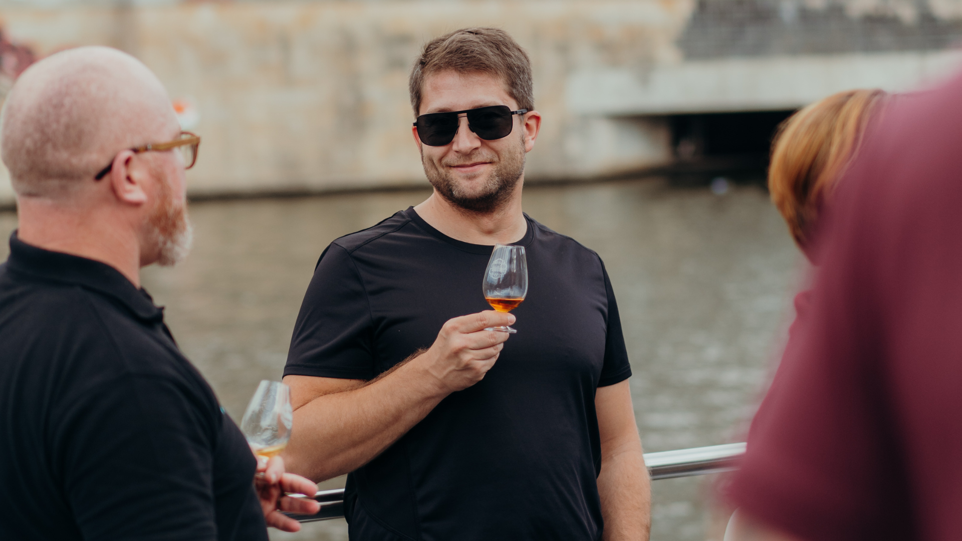 Man with dark sunglasses holding a filled whisky glass, standing on a boat. There are 2 people beside him.