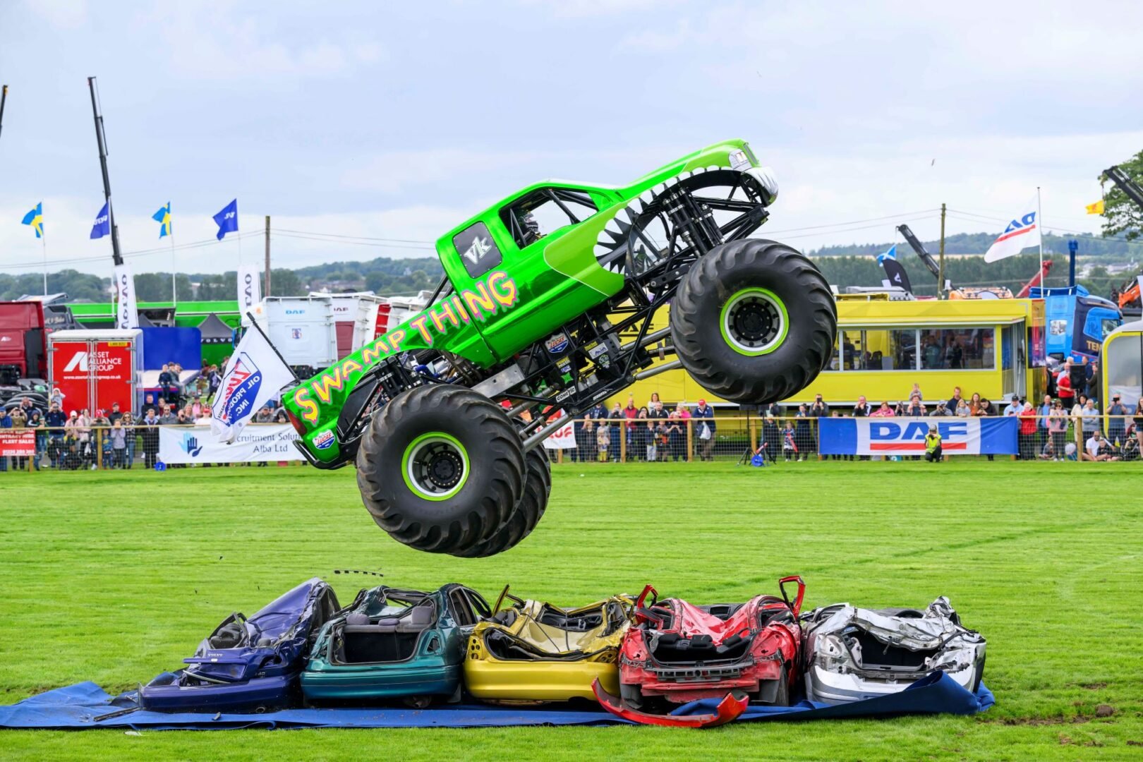 Large green truck driving over 5 squashed cars.