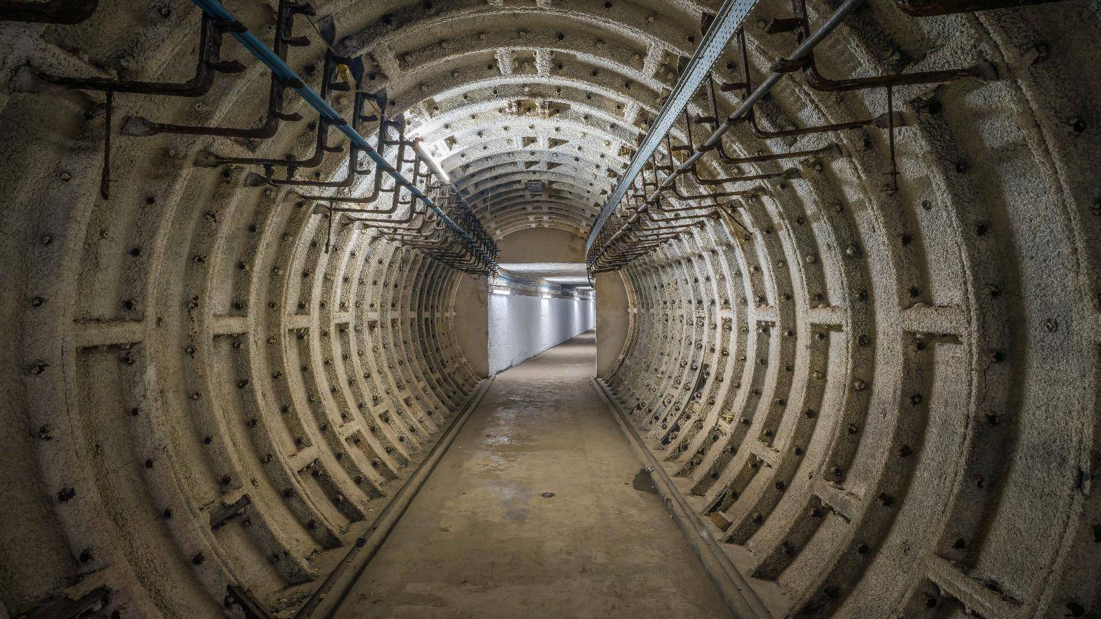 A rounded metal tunnel lined with pipes, leading to a narrow corridor