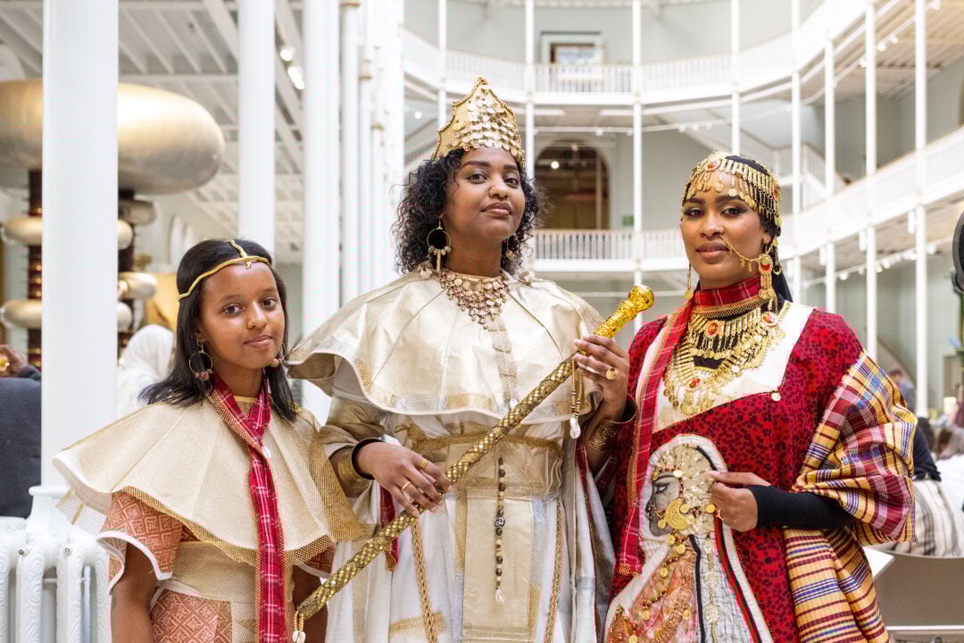 Two women and a girl wearing traditional costumes and gold headpieces. The woman in the middle, in a cream and gold outfit, carries a gold sceptre.