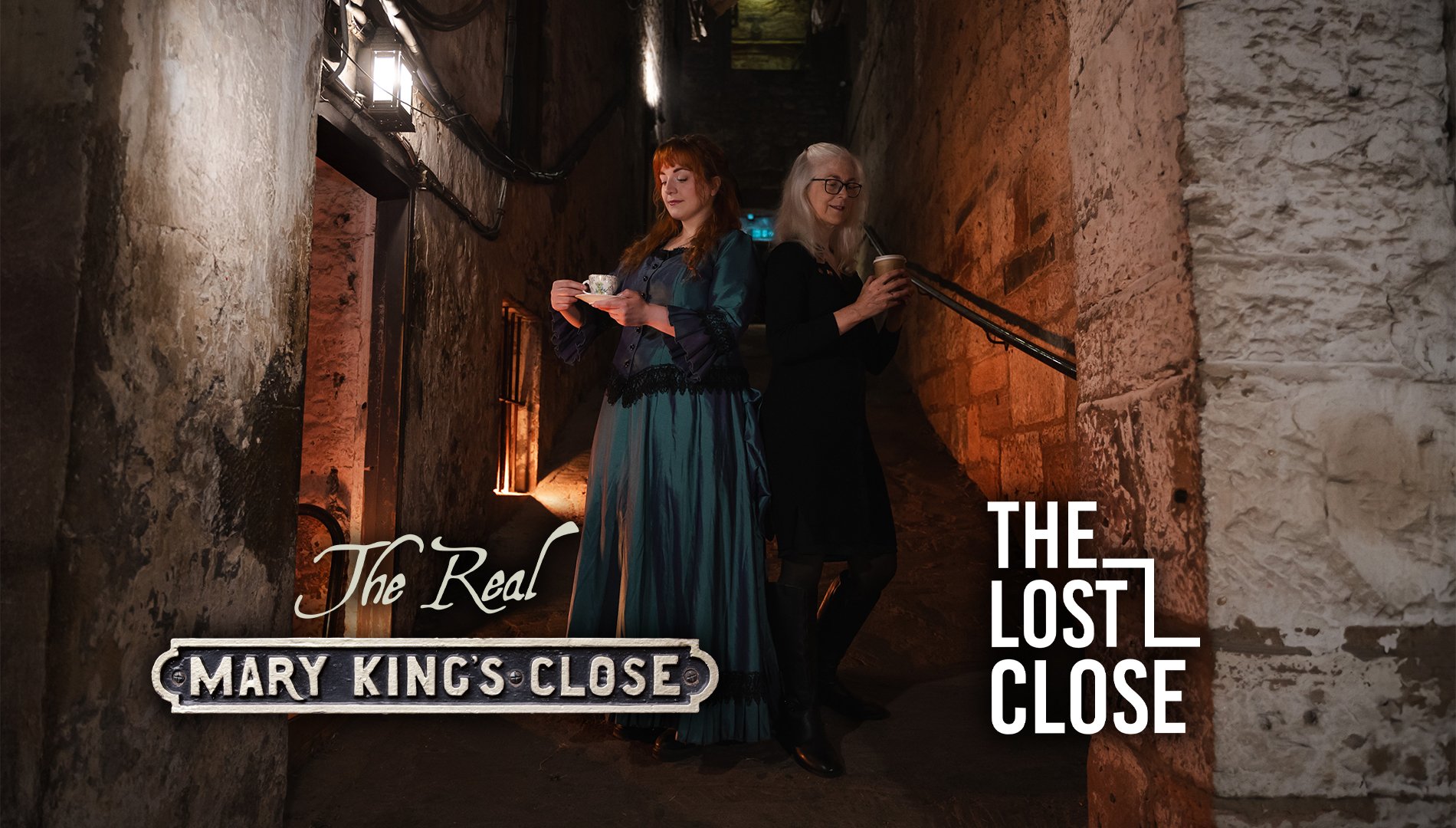 2 women, one wearing a long, old fashioned dress, standing in an underground close. One holds a teacup and saucer, the other a paper coffee cup.