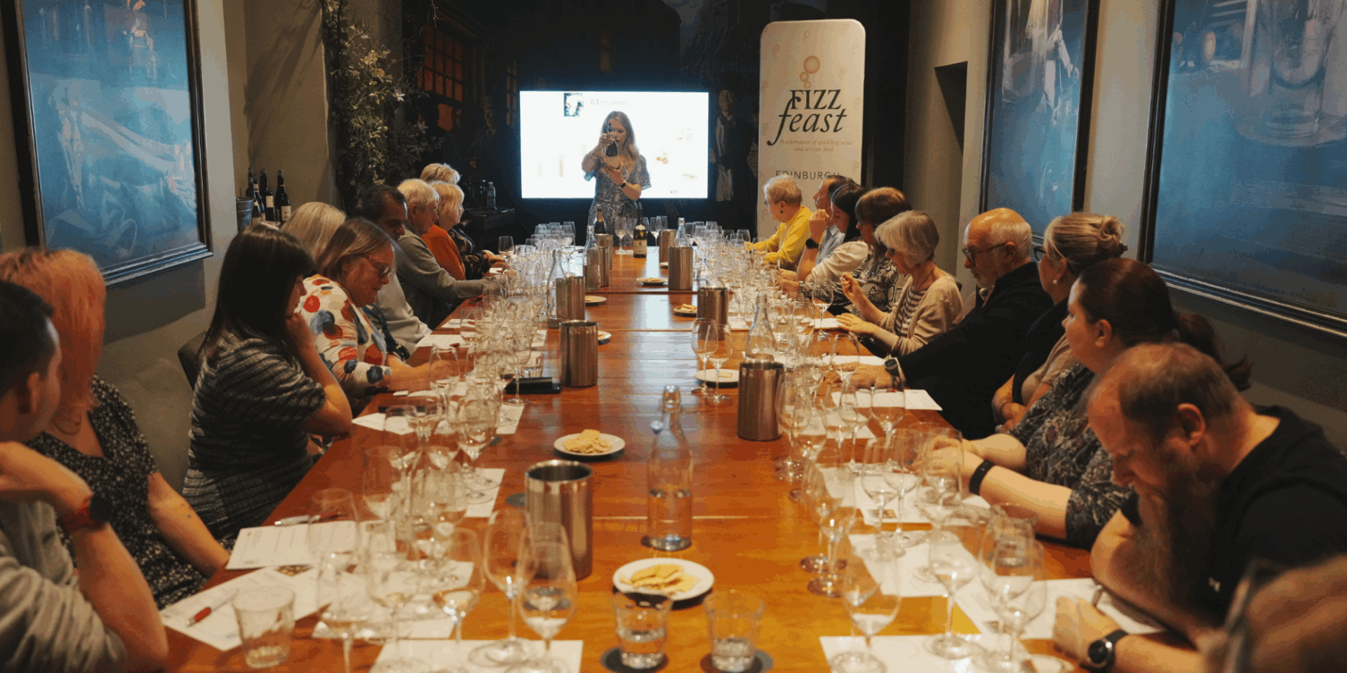 Room full of people, sitting at a long table, with a women standing at the top of the table. In front of each person are a selection of wine glasses.