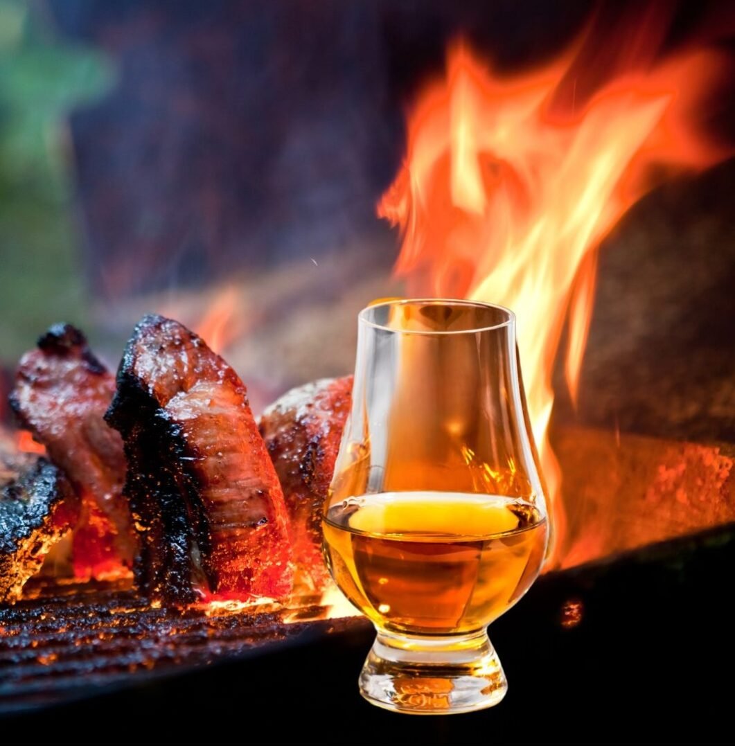 Small glass of whisky, sitting in front of a flaming BBQ, which has meat steaks cooking on it.