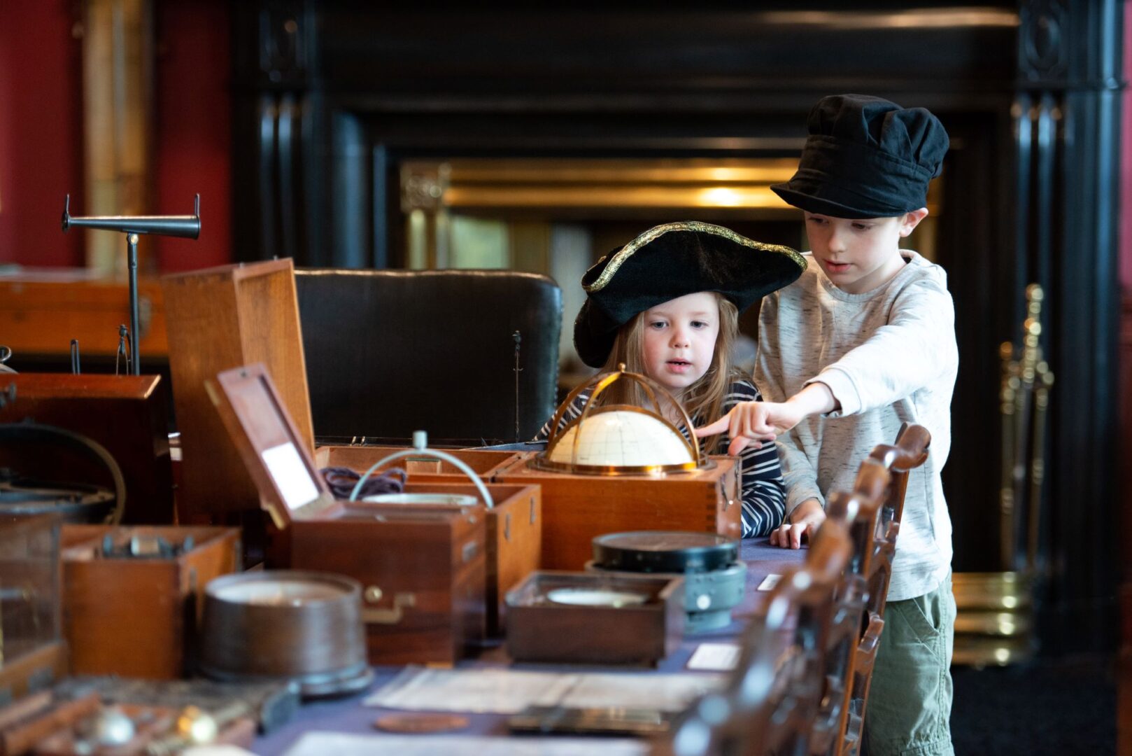 A young boy and girl, both wearing old fashioned hats, look at an assortment of old objects on a table.