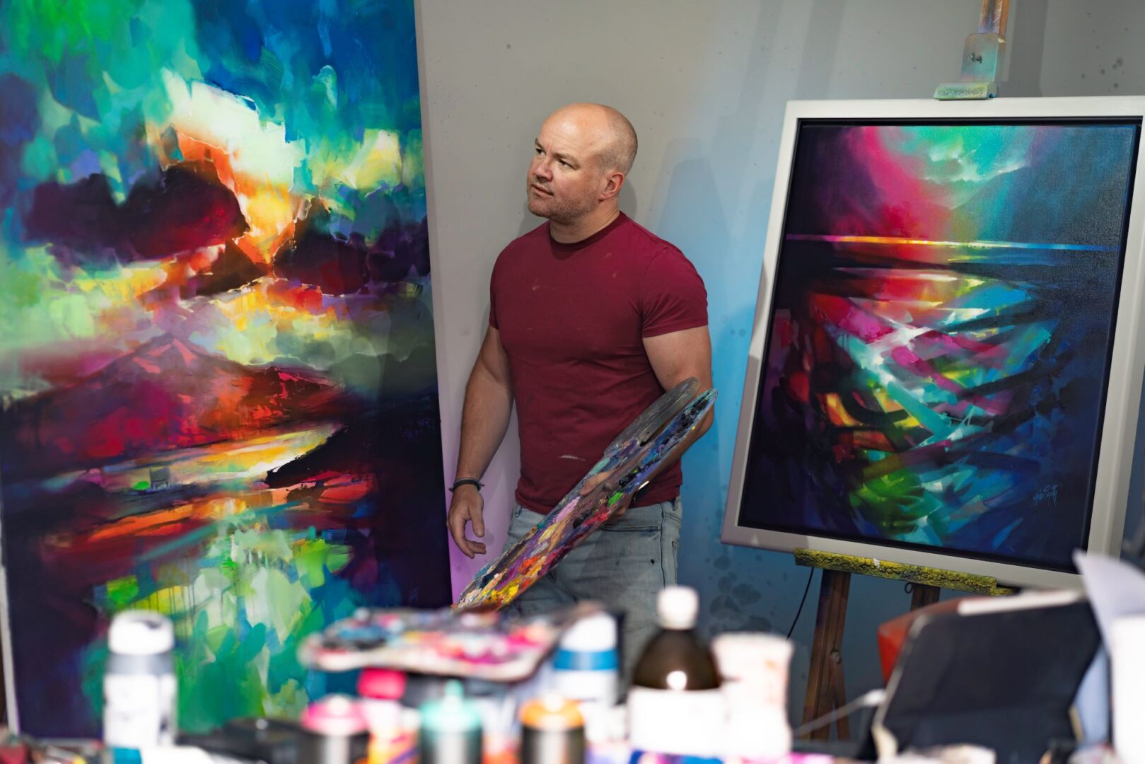 Man standing with an artist's painting board, surrounded by colourful canvases and bottles of paint.