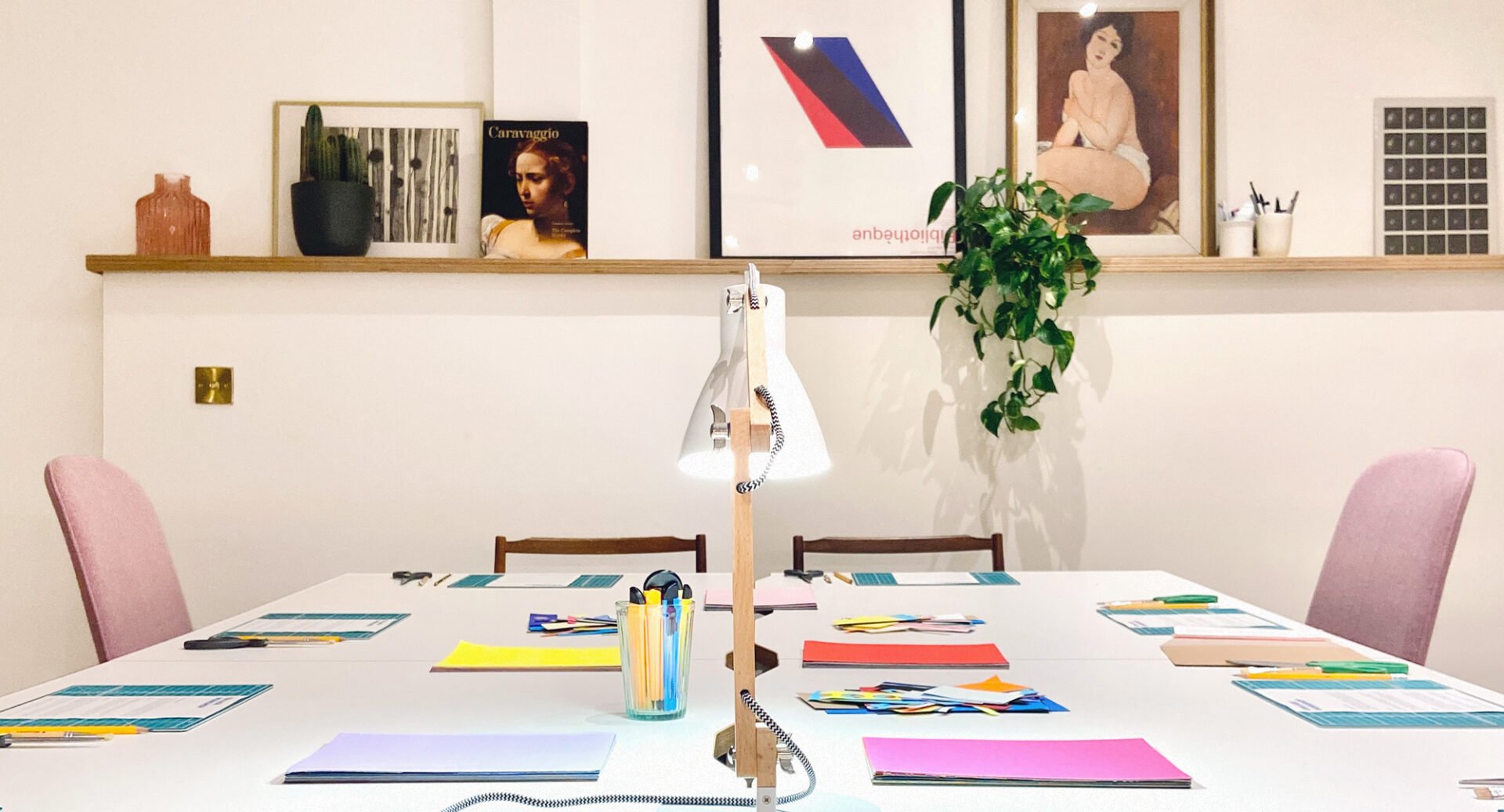 A table, set with piles of coloured papers, a lamp and a tub of pens and pencils.