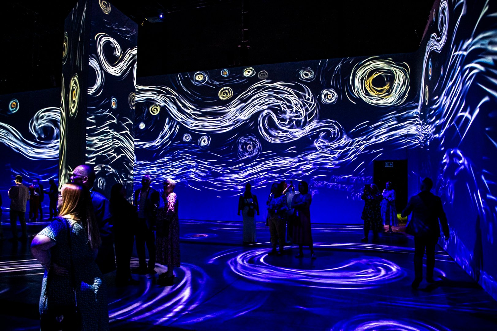 People standing in darkened room, the walls of which have projections of white swirls on a dark blue background