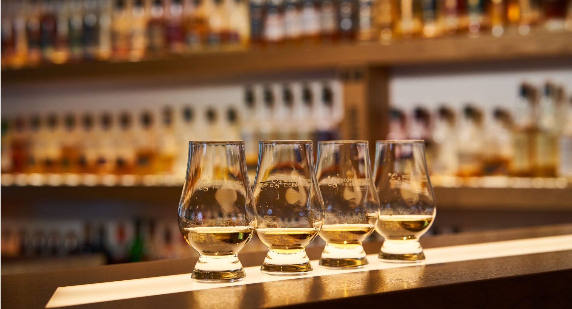 A row of four glasses containing whisky, with the blurred background of whisky bottles on the bar