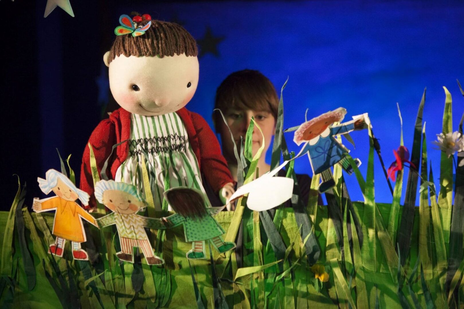 A toy doll sitting behind a row of cut-out people. In the background can be seen a women's head, looking at the scene.