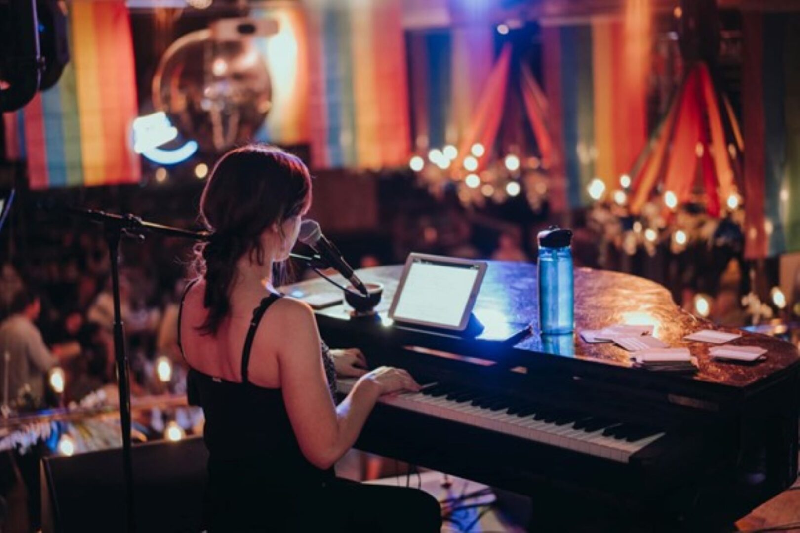 A side-view of a person sitting at a piano, playing in front of a crowded room.