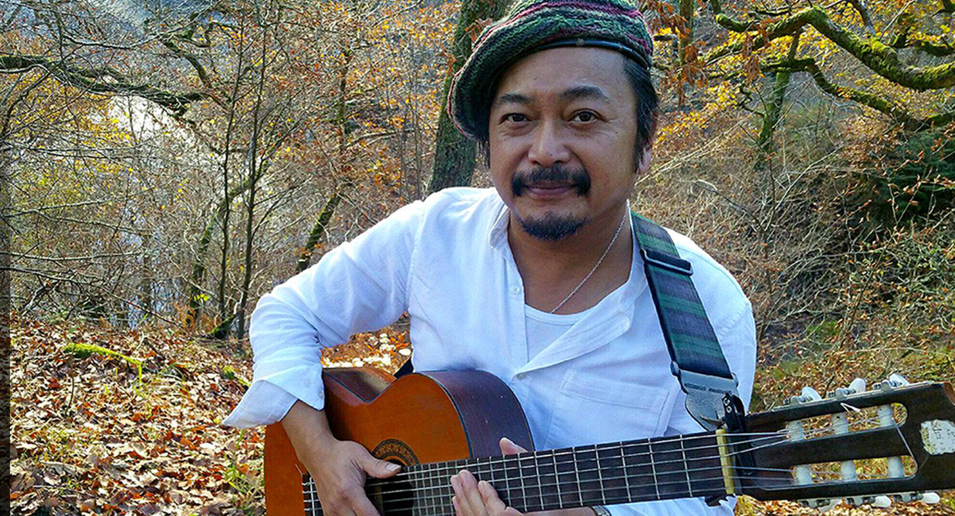 Man sitting in a wooded area, holding a guitar.