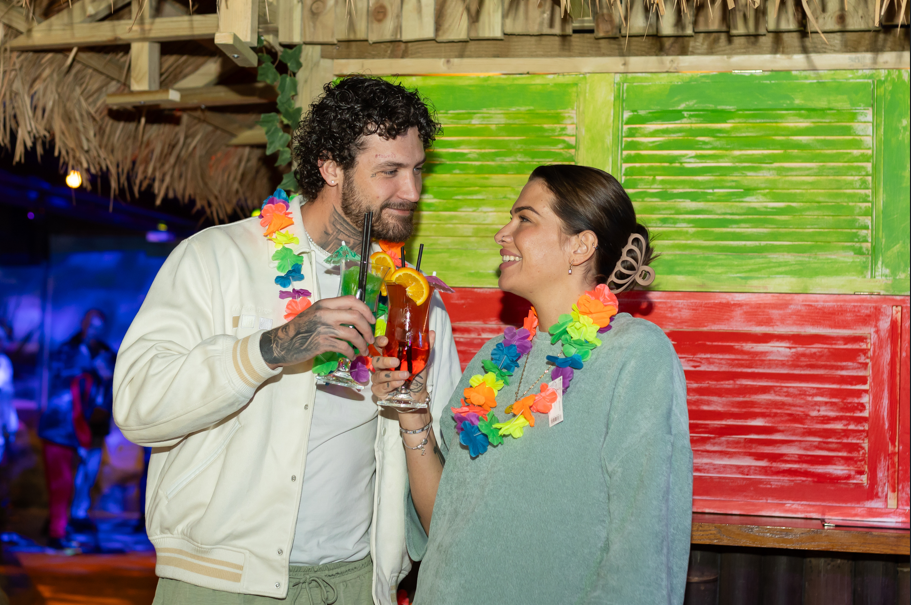 A woman and man wearing hawaiian flowers around their neck smiling and holding drinks