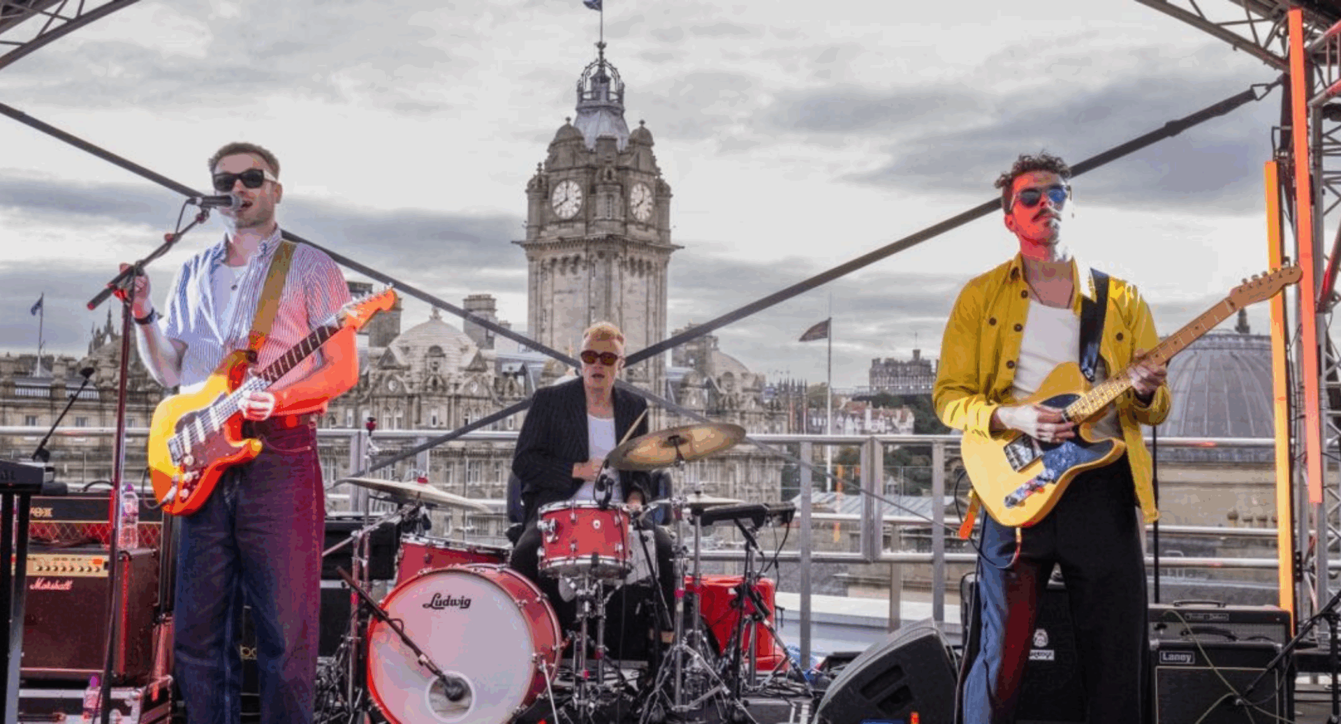 Band, comprising 2 guitarists and a drummer performing outdoors, against the backdrop of a tall clock tower.