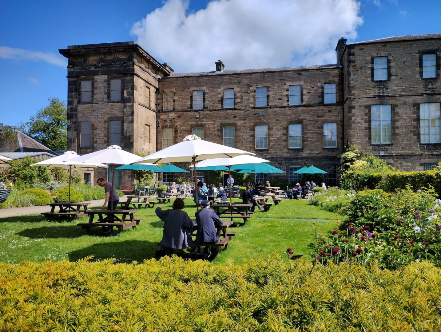 Groups of people sitting outdoors on benches. In the background is a large stone building.