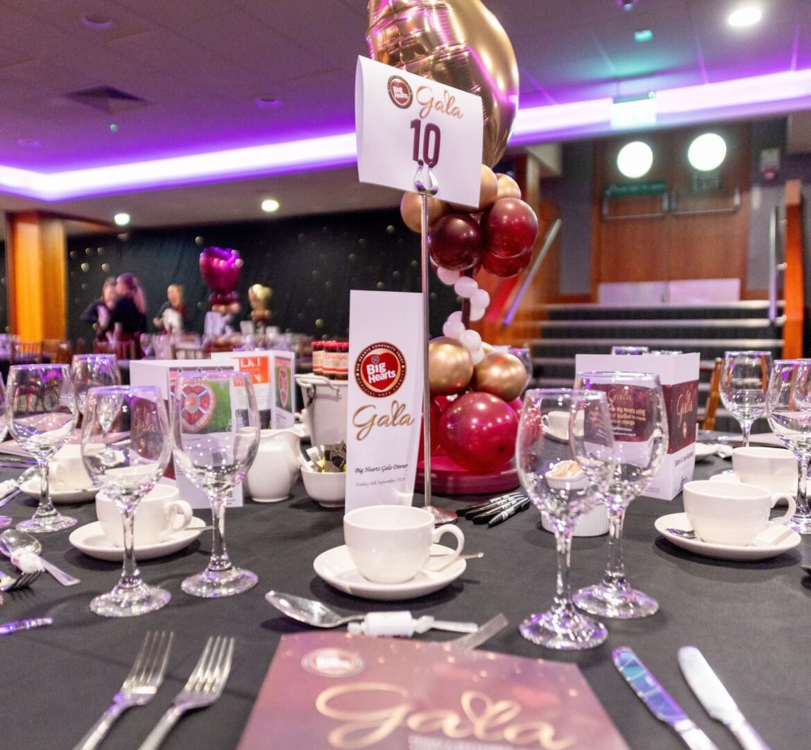 A table set with wine glasses, cups and saucers and cutlery. In the middle is a large display of red and gold balloons.