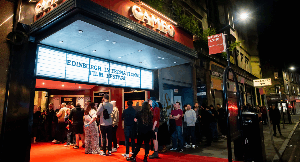 Night-time image of a queue of people waiting outside a brightly lit cinema building.