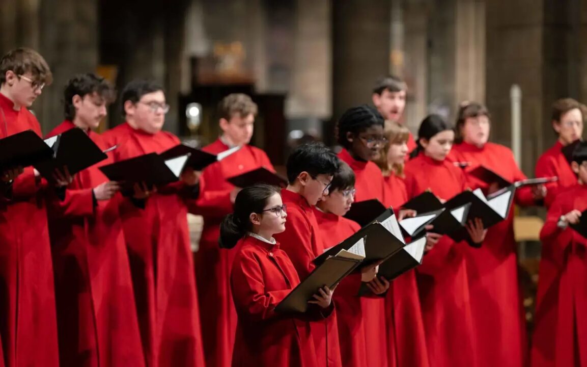 A group of adults and children, dressed in long red cloaks and holding a large book, singing.