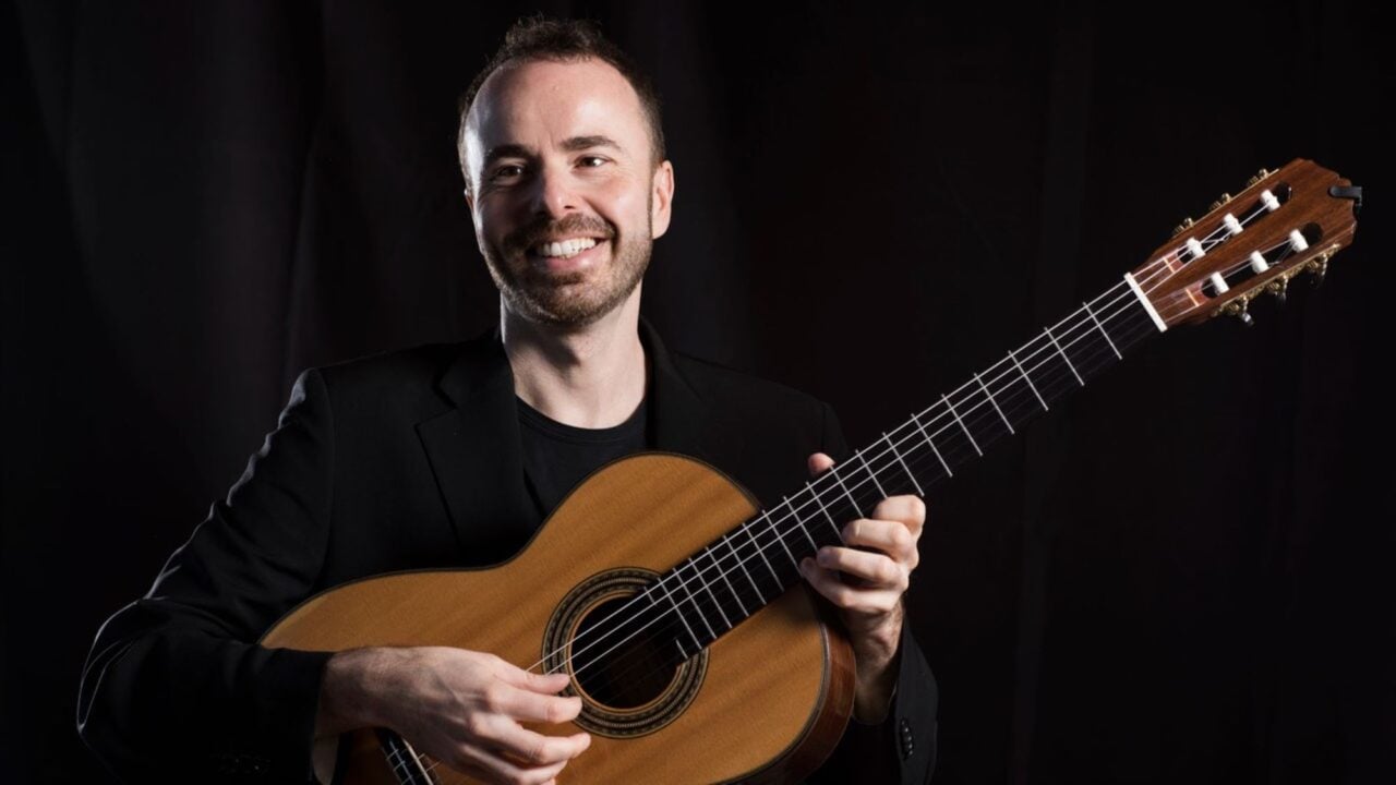 Man against a plain black background, and wearing a black jacket, holding a guitar.