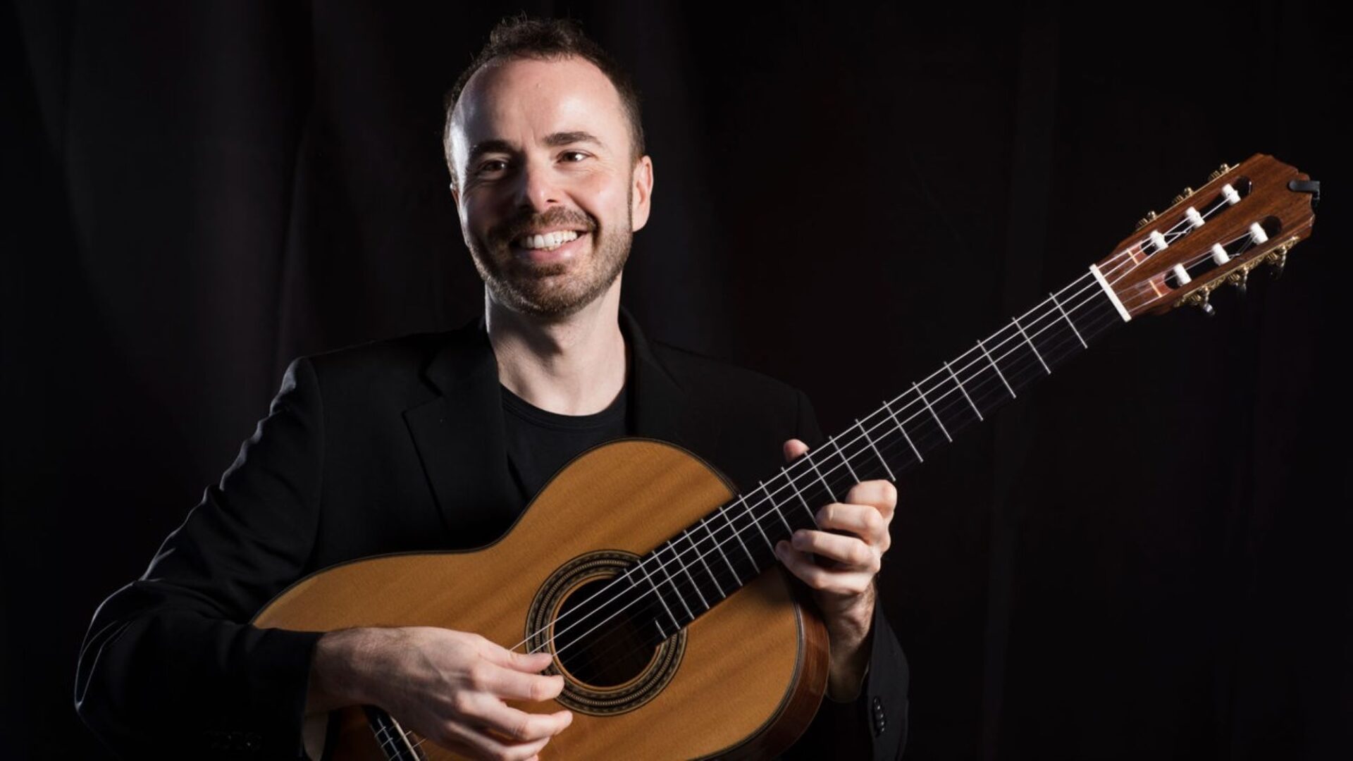 Man against a plain black background, and wearing a black jacket, holding a guitar.