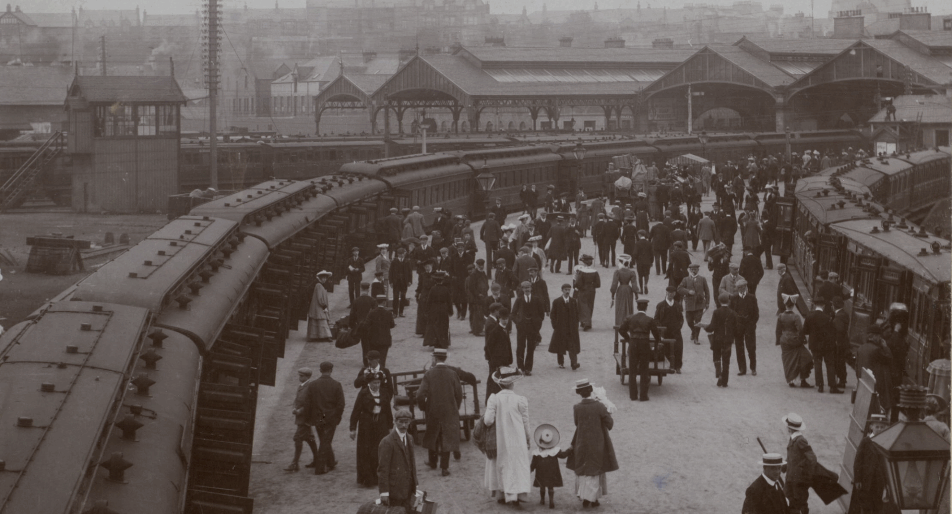 Black and white photo of a 1900s train station platform, on which there are crowds of people boarding trains.