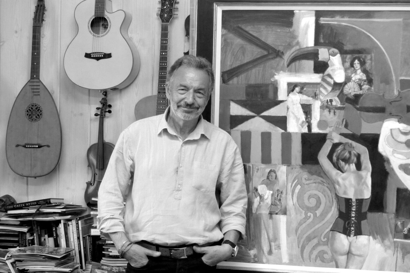 Black and white image of a man standing against a wall, on which hangs various musical instruments and a large painting of several female figures.