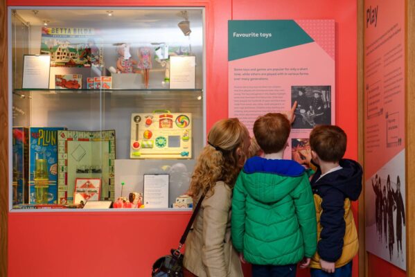 Image shows some favourite classic toys in a glass case and a mother reading the description to her two sons