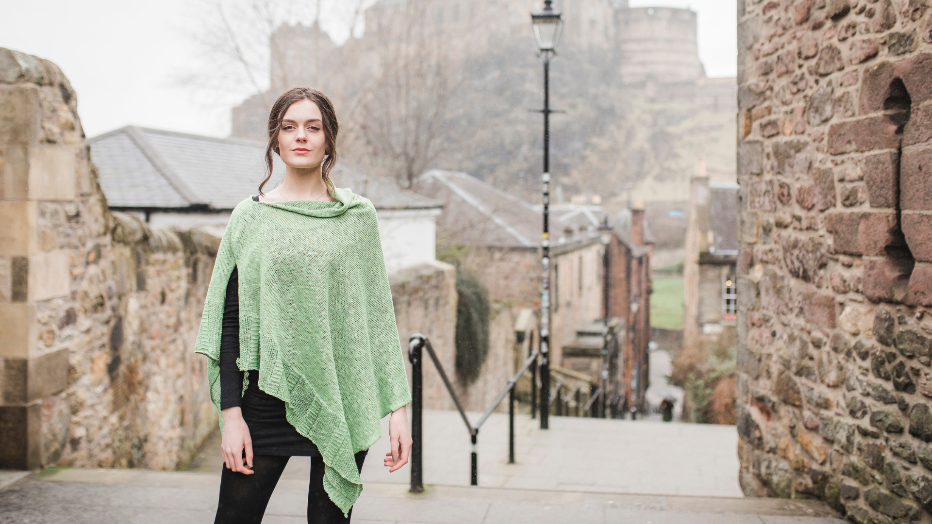 A woman wearing a Bill Baber Knitwear shawl at the top of stairs in front of Edinburgh Castle at the Edinburgh Vennel,© Bill Baber Knitwear
