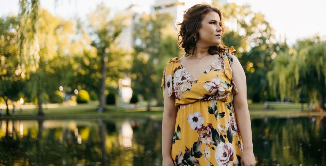 A women wearing a yellow flowery dress standing in front of a group of large trees.