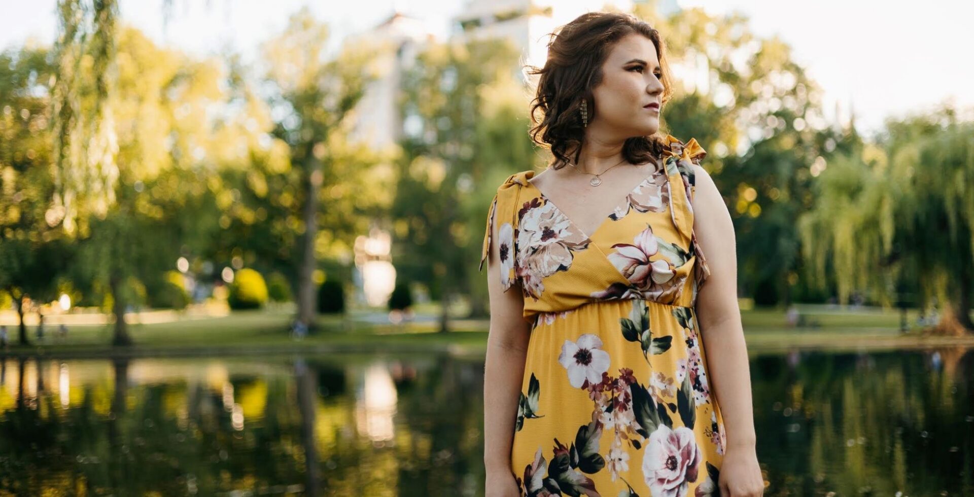 A women wearing a yellow flowery dress standing in front of a group of large trees.