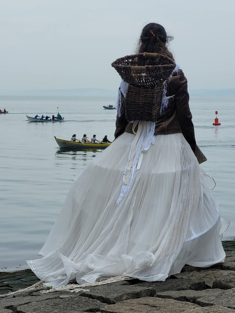 A woman in traditional fisherwoman garb with a creel on her back looks out over the Forth and the skiffs on the water