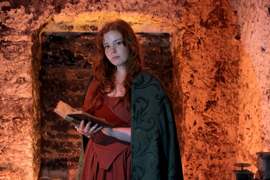Woman dressed in old fashioned clothing holding a large book. She is standing in an underground room.