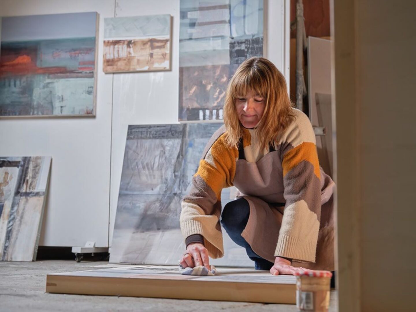 A woman kneeling over a painted canvas. On the wall behind her are several paintings.