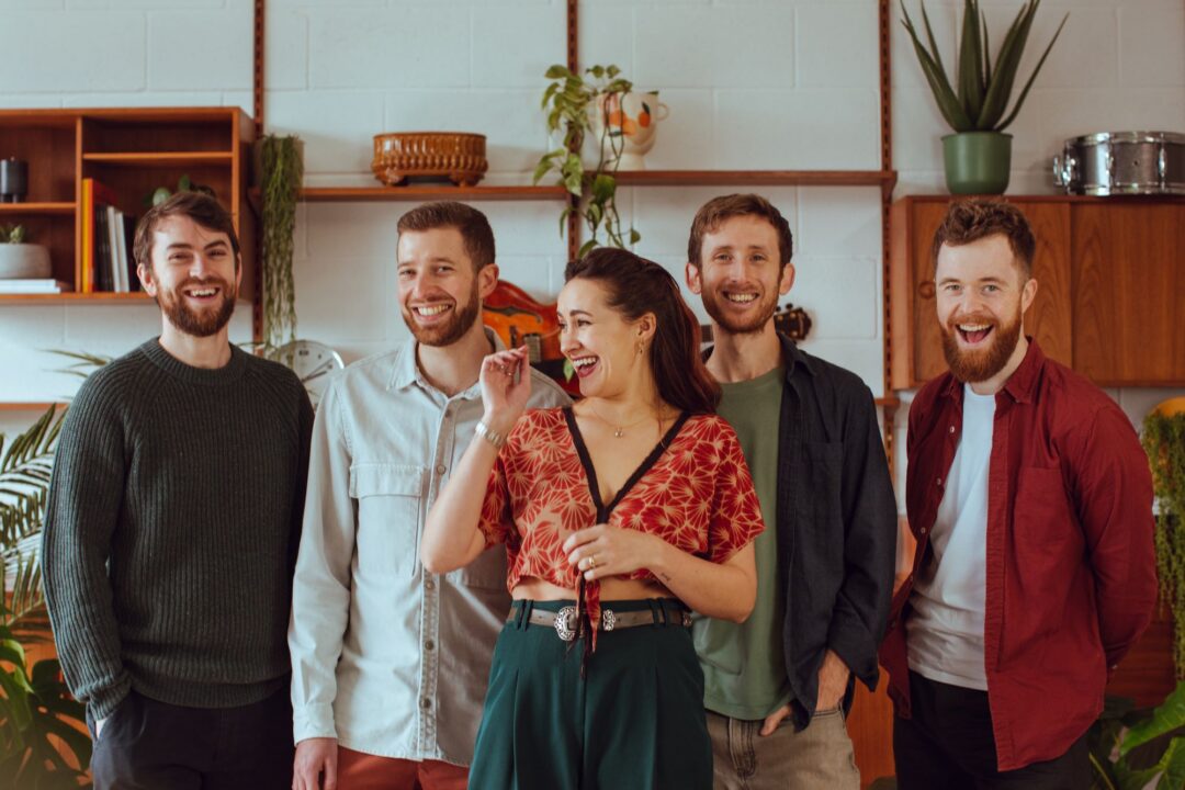 Group of 4 men, with a woman stood in front of them. Behind them are walled shelf units, decorated with plants.