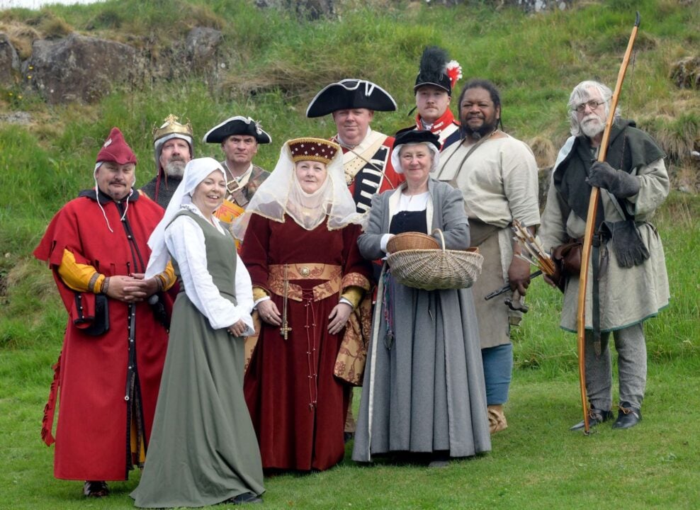 A group of people dressed in old fashioned dress, against a grassy hill.