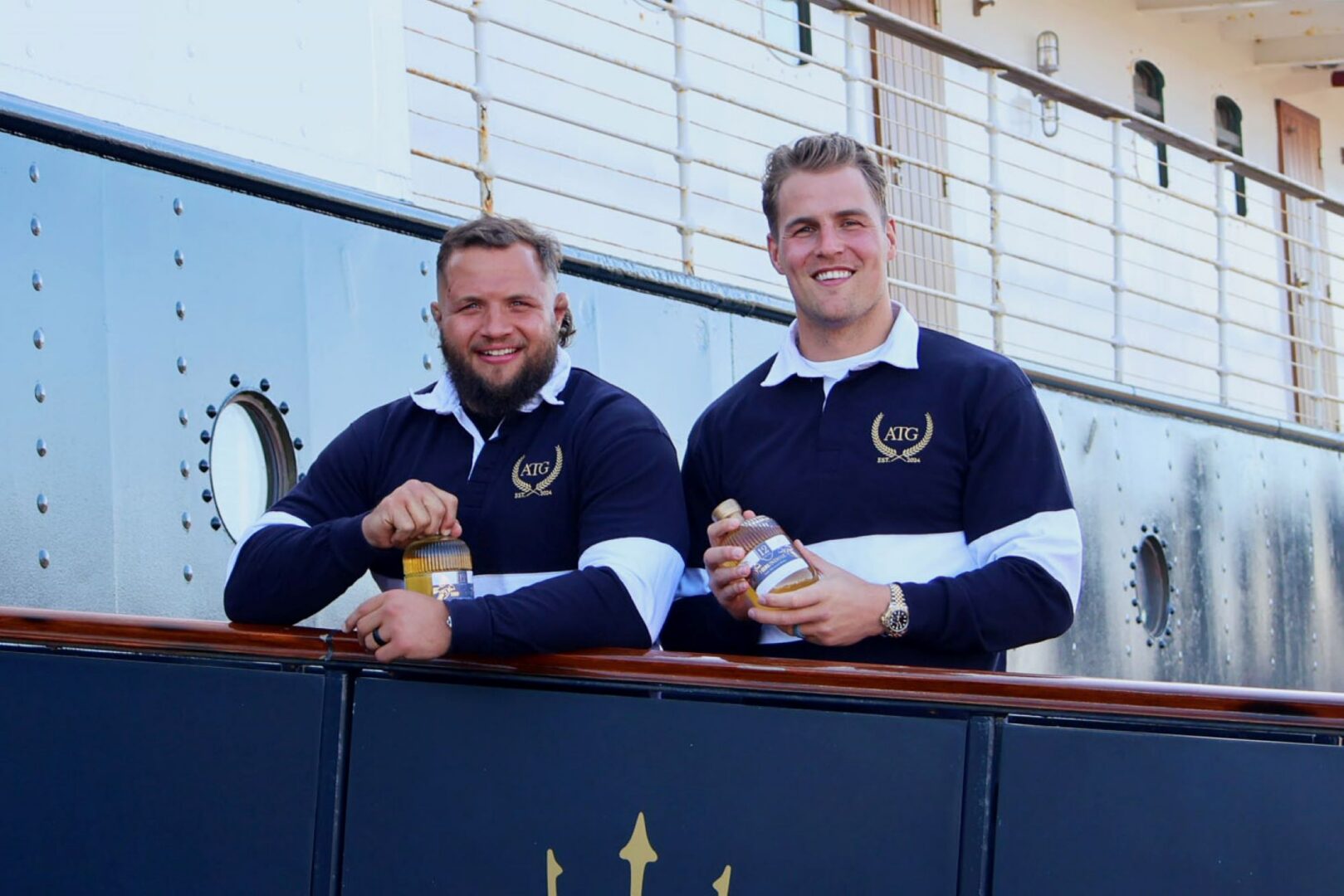 2 men standing on the deck of a ship, each holding a small bottle of whisky.