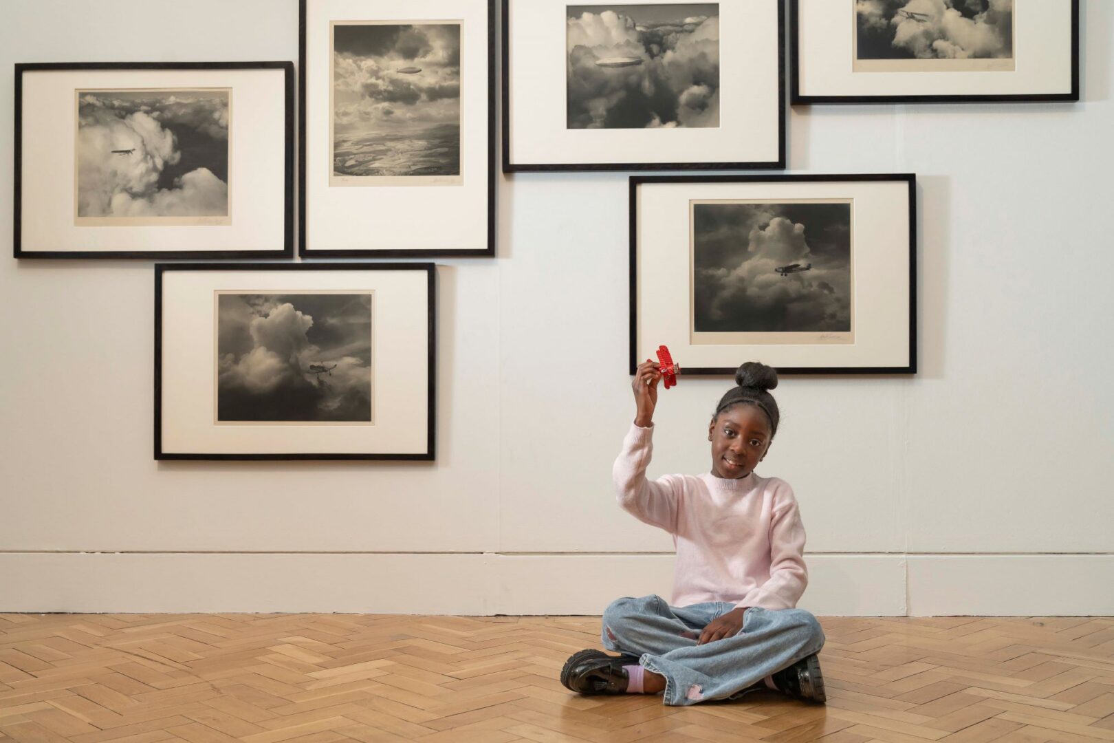 Young girl sitting cross-legged on the floor, in from of a wall of framed photos.