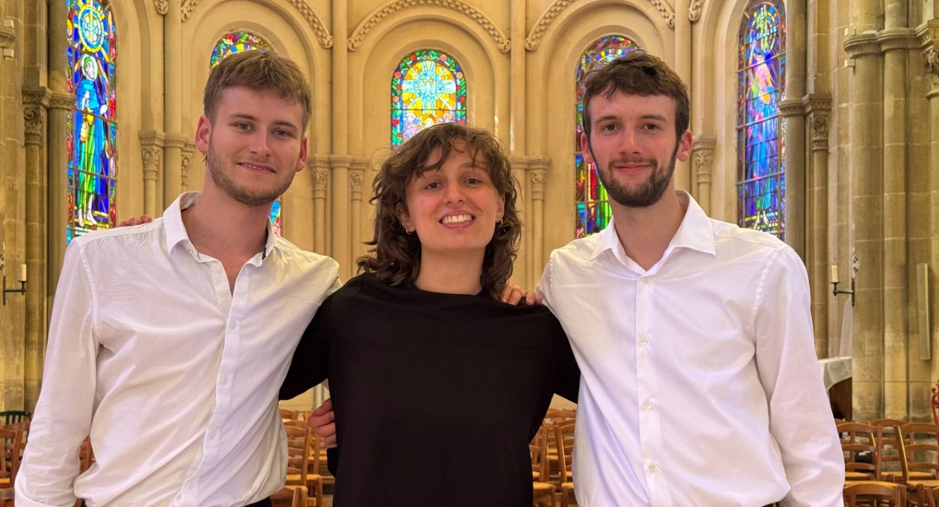 3 people standing against a wall , with bright stained-glass windows.