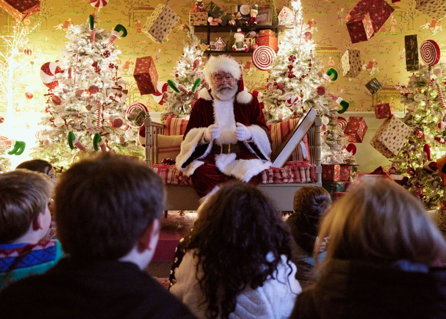 A man dressed as Santa sitting on a bench, surrounded by Christmas trees and wrapped presents. In the foreground, young children sit on the floor facing him.