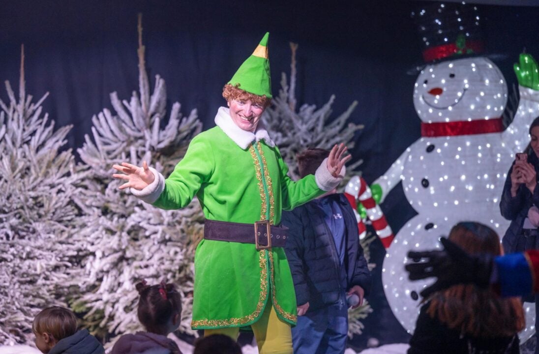 A man dressed in a green elf costume, standing in front of a snowy tree and a giant snowman.