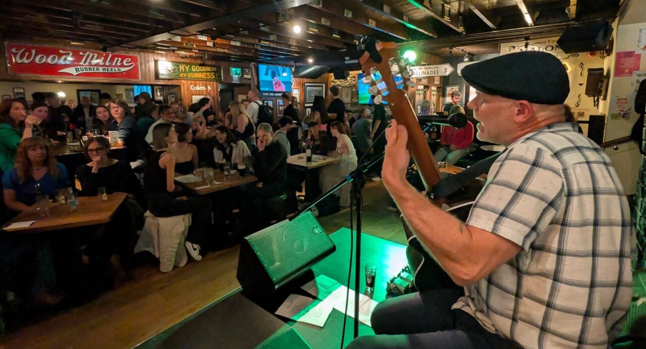 Man playing a guitar and singing on stage in front of a bar filled with people.