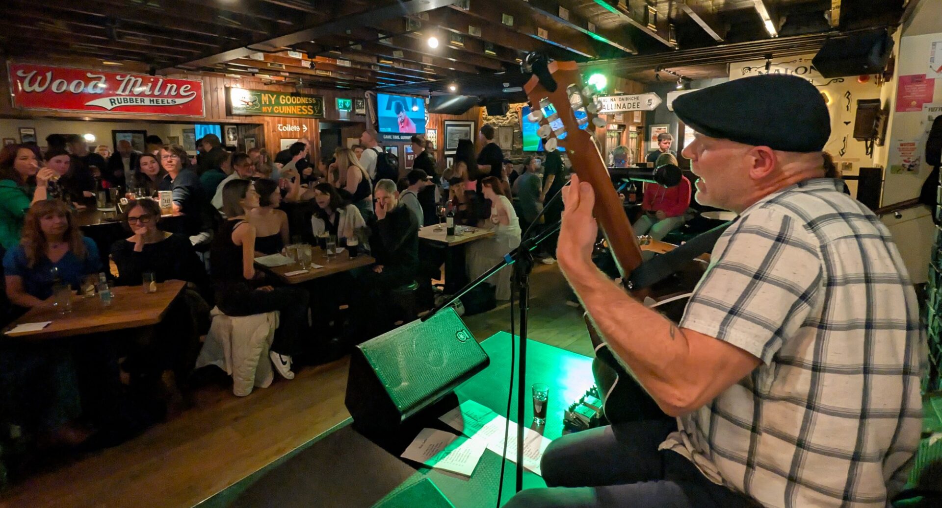Man playing a guitar and singing on stage in front of a bar filled with people.