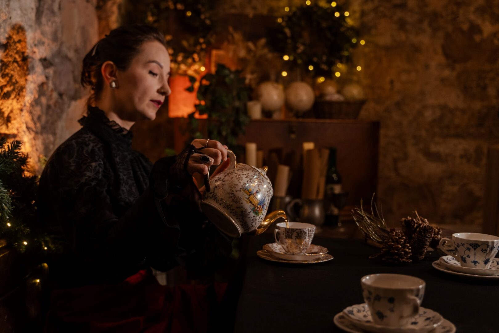 A woman wearing old fashioned dress, sitting down, pouring tea from a pot into a cup and saucer.