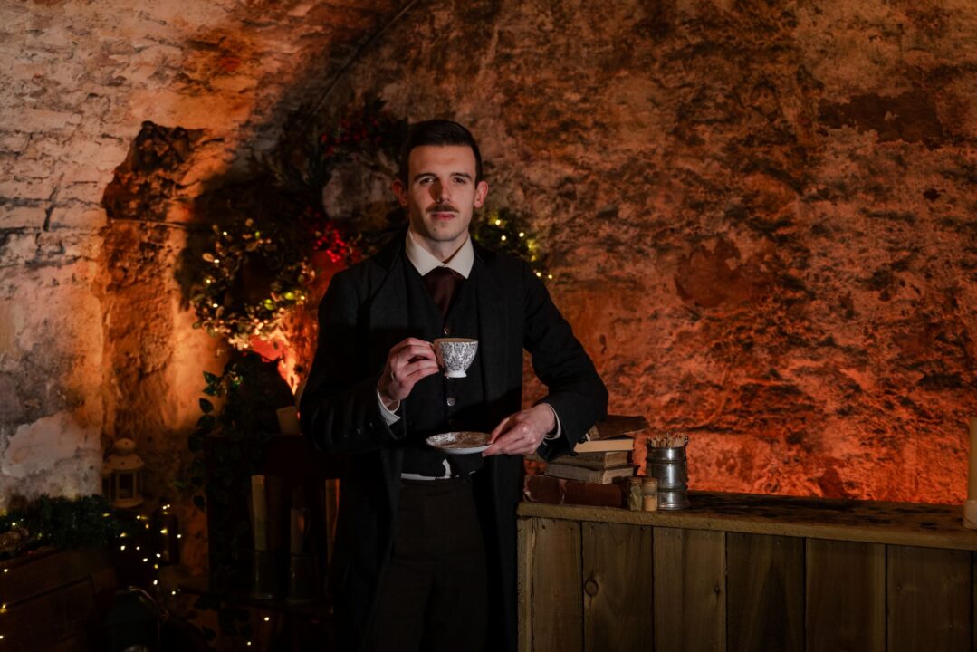 A man dressed in old fashioned clothing holding a cup and saucer. Behind him is a light walled cavern.