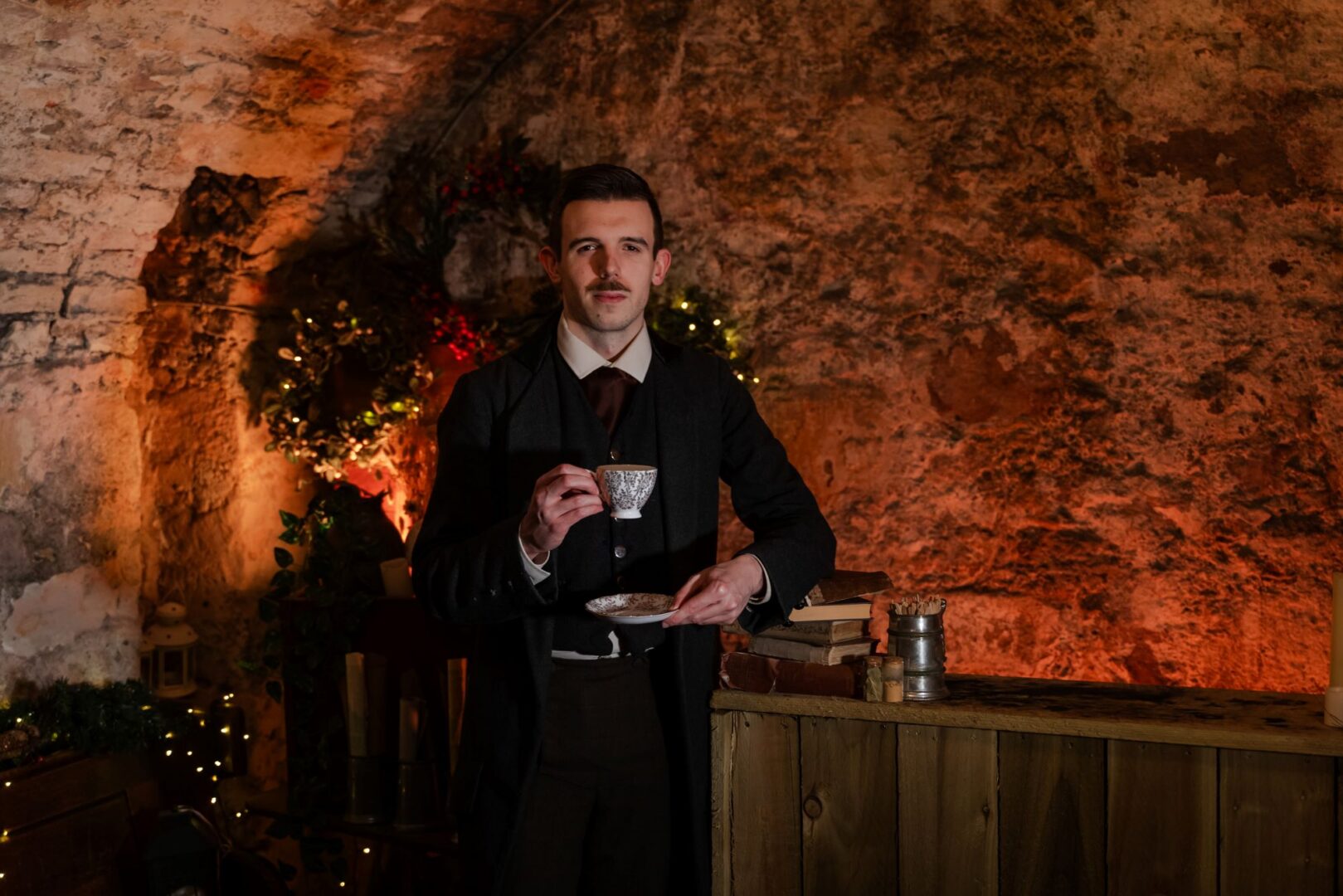 A man dressed in old fashioned clothing holding a cup and saucer. Behind him is a light walled cavern.