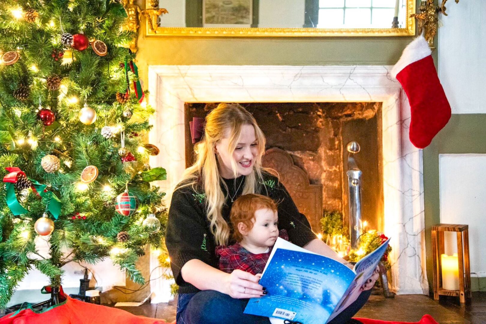 Woman sitting on floor with young girl on her knee, looking at a large book. Behind them is a decorated Christmas tree and fireplace.