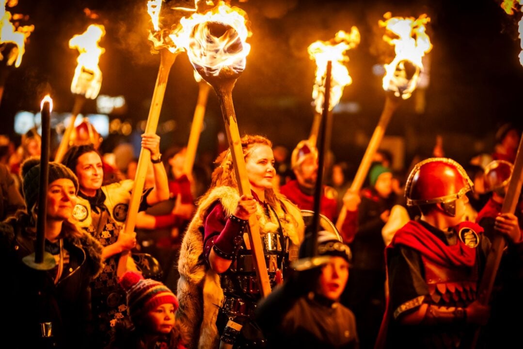 A group of whom marching through a street at night. Some are dressed in traditional Viking clothing and holding firelit torches.